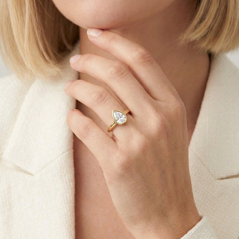 May include: A gold ring featuring a pear-shaped, clear gemstone. The ring has a thin, polished gold band and a bezel setting that surrounds the gemstone. The ring is displayed on a person's finger.