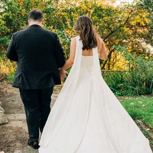 May include: A black suit jacket and matching trousers paired with a floor-length white wedding dress featuring a corset-style lace-up back, a long flowing skirt, and a row of small buttons running down the centre of the back of the gown.
