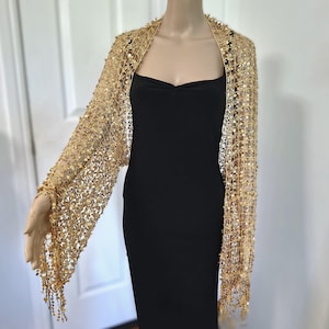 May include: A gold sequin shawl with long sleeves and a draped design. The shawl is worn over a black dress. The sleeves have a fringe detail at the wrist. The shawl is made of a delicate, open-weave fabric.
