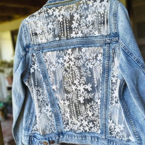 May include: Pale blue denim jacket with a white floral lace back panel. The jacket has a classic collar, button fastenings, and pockets. The back features a delicate, see-through lace design with floral patterns.