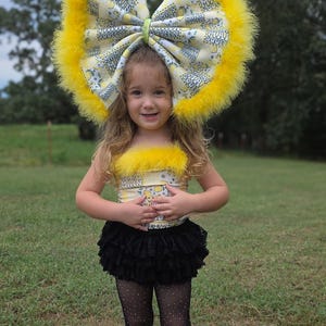 Puede incluir: Una niña pequeña con un atuendo de temática de limón amarillo y blanco, que incluye un lazo grande, una parte superior y calentadores de piernas. Lleva pantalones cortos negros y medias de rejilla. El atuendo está acentuado con plumas amarillas. Está de pie sobre hierba verde.