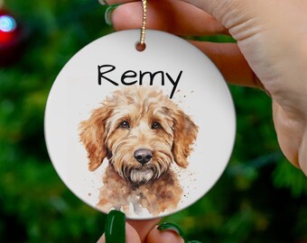 Peut inclure: Une boule de Noël en céramique blanche avec une peinture à l'aquarelle d'un chien brun et blanc. La boule a un cordon doré et le nom "Remy" écrit en noir.