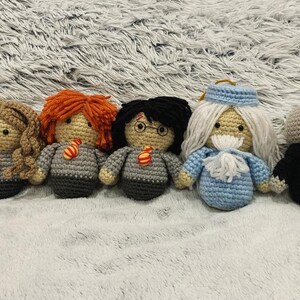 May include: A set of seven crocheted Harry Potter characters, including Harry Potter, Hermione Granger, Ron Weasley, Albus Dumbledore, Severus Snape, and a Dementor. The characters are all wearing their Hogwarts uniforms and are standing on a fluffy white surface.