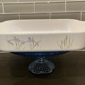 Puede incluir: Una fuente rectangular blanca con asas, decorada con flores de iris moradas y tallos verdes, se encuentra sobre un soporte para pasteles de vidrio azul. El plato es probablemente de cerámica o porcelana.