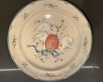 May include: A beige ceramic dinner plate with a blue rim and a floral design featuring three white birds with yellow beaks and a red apple. The birds are holding a blue ribbon.