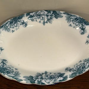 May include: Oval serving platter with a white center and a teal floral border. The flowers are detailed and arranged around the edge of the platter. The rim has a scalloped edge and a gold trim. A decorative piece for serving food.
