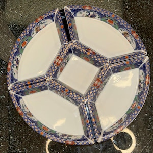 Peut inclure: Un plat de service en céramique blanche avec un motif floral bleu et doré. Le plat est divisé en six sections, avec un carré central et cinq compartiments en forme de tarte. Le bord présente un motif détaillé avec des éléments floraux et géométriques.