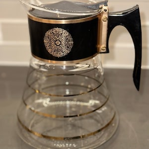 May include: A vintage glass coffee jug with a black and gold design. The jug has a clear glass body with gold horizontal stripes and a black top with a gold decorative emblem. The handle is black and the lid is clear.