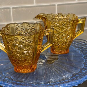 May include: Amber glass sugar bowl and creamer set with geometric starburst pattern. The set has handles and sits on a blue glass serving plate. The creamer has a spout. The glass has a warm, golden hue.
