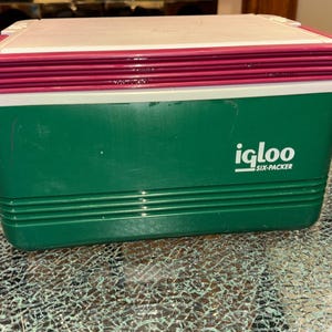 Op de afbeelding: Een groene Igloo Six-Packer koeler met een witte deksel en een magenta rand. De koeler heeft een rechthoekige vorm met horizontale ribbels en het Igloo-logo aan de zijkant. Hij staat op een glazen tafel.