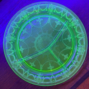 May include: A round, divided glass plate glowing with a vibrant neon green hue under ultraviolet light. The surface features an intricate, etched pattern of swags, garlands, and central radial motifs, creating a detailed decorative border.