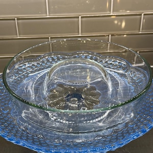 Może przedstawiać: Szklany stojak na ciasto z przezroczystego szkła z niebieską podstawą i falistym brzegiem. Stojak ma zdejmowaną górną miskę.