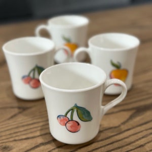 May include: Four white ceramic mugs with fruit designs. Two mugs feature cherries, one mug features an orange, and one mug features an apple.
