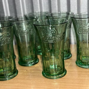 Könnte beinhalten: Ein Satz von acht grünen Glasbechern mit dem Coca-Cola-Logo, das auf der Seite eingeprägt ist. Die Gläser sind klar und haben eine leicht gefrostete Optik.