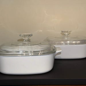 May include: Two white casserole dishes with clear glass lids. The dishes are rectangular with rounded corners and have small handles on each side. The lids have a clear, decorative knob. The dishes are on a dark surface.