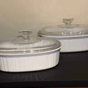 May include: Two oval white ceramic baking dishes with vertical ribbed sides and clear glass lids. Each lid features a central knob handle. The dishes are arranged with one positioned slightly behind and to the right of the other on a flat surface.