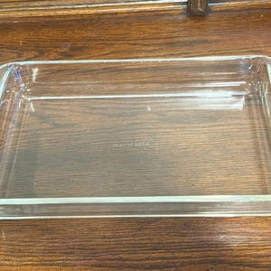 Könnte beinhalten: Eine rechteckige Backform aus klarem Glas mit abgerundeten Ecken. Die Form ist leer und steht auf einer Holzoberfläche. Die Form hat ein schlichtes, klassisches Design, geeignet zum Backen und Servieren.