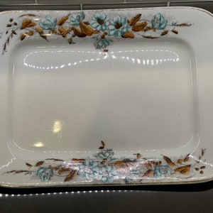 May include: A white rectangular serving platter with a gold rim and a floral design in shades of brown, blue, and green.