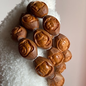May include: A brown wooden bracelet featuring carved animal faces. The bracelet is made of oval-shaped beads, each with a different animal design. The beads are strung together with a clear, elastic cord. The bracelet is displayed on a white, fluffy surface.