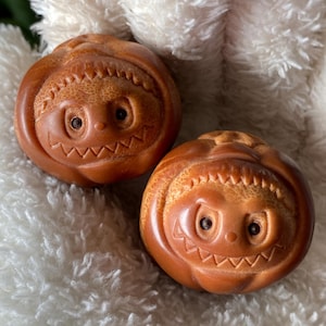 May include: Two small, round, wooden carvings with smiling faces. The carvings are a warm brown color and have detailed features, including eyes, a mouth, and a textured top. They rest on a fluffy white surface.