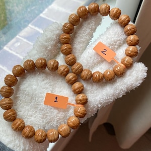 May include: Two bracelets made of light brown, carved beads. Each bead has a detailed, textured design. The bracelets are displayed on a white, fluffy surface. Orange tags labeled "1" and "2" are attached to the bracelets.