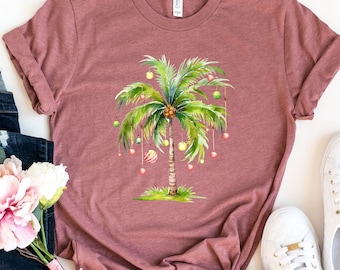 May include: A mauve t-shirt featuring a graphic of a palm tree adorned with Christmas baubles. The t-shirt is laid flat on a white background with a pair of white trainers and a pair of blue jeans with a bunch of pink flowers.