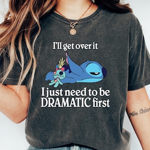 Op de afbeelding: Een donkergrijs T-shirt met een cartoonfiguur van Stitch en de tekst "I'll get over it I just need to be DRAMATIC first".