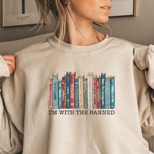 Może przedstawiać: Beżowa bluza z nadrukiem rzędu starych książek z tekstem "I'M WITH THE BANNED" wydrukowanym poniżej.