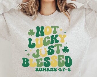 May include: A light grey sweatshirt with the text "Not Lucky Just Blessed" in green, yellow, and white letters with shamrock designs. The text "Romans 4:7-8" is printed below the main text.