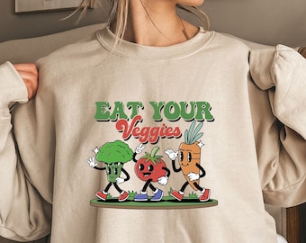 May include: A beige sweatshirt with the text "Eat Your Veggies" and a cartoon illustration of a broccoli, tomato, and carrot walking.