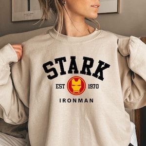Könnte beinhalten: Beigefarbenes Sweatshirt mit Rundhalsausschnitt und dem Wort "STARK" in schwarzen Blockbuchstaben. Darunter befindet sich ein rot-gelbes Iron Man-Logo mit den Worten "EST 1970" darüber und "IRONMAN" darunter. Das Sweatshirt hat eine neutrale Farbe.