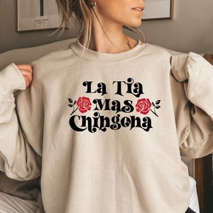 Op de afbeelding: Een beige sweatshirt met de tekst "La Tia Mas Chingona" in zwart met rode rozen aan weerszijden van de tekst.