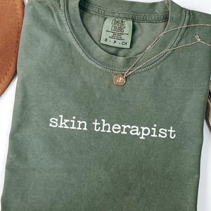 May include: An olive green t-shirt with the words "skin therapist" printed in white. A gold necklace with a small pendant rests on the shirt. A brown hat and dried palm leaves are in the background.