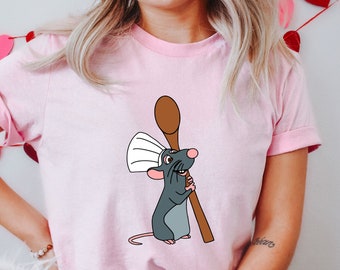 May include: A pink t-shirt featuring a cartoon rat wearing a chef's hat and holding a wooden spoon. The rat is grey and white and has a friendly expression.