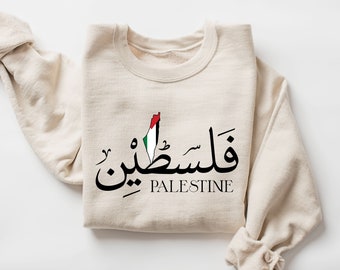 Op de afbeelding: Een beige sweatshirt met het woord "Palestina" in Arabisch schrift en Engelse tekst. Het ontwerp bevat een kaart van Palestina met de kleuren van de Palestijnse vlag. De mouwen zijn opgerold.