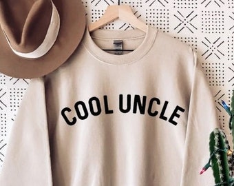 May include: A beige sweatshirt with the text "COOL UNCLE" printed in black on the front. The sweatshirt is hanging on a hanger against a white background with a black geometric pattern.