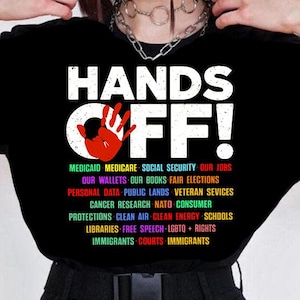 May include: Black t-shirt with the text "HANDS OFF!" in white with a red handprint. The text below the handprint lists various social and political issues in rainbow colors.
