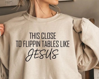 May include: A beige sweatshirt with the text "This close to flipping tables like Jesus" printed in black.