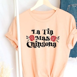 May include: Peach colored t-shirt with black text that reads "La Tia Mas Chingona" with a red rose graphic.