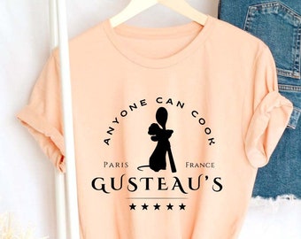 May include: A peach-coloured t-shirt with a black graphic that says "Anyone can cook" with a silhouette of a rat holding a spoon. The graphic also includes the words "Paris" and "France" and the text "Gusteau's" with four stars.