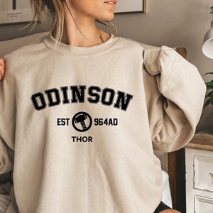 Op de afbeelding: Een beige sweatshirt met zwarte tekst die "ODINSON EST 964AD THOR" luidt met een zwarte cirkel met een hamerafbeelding.