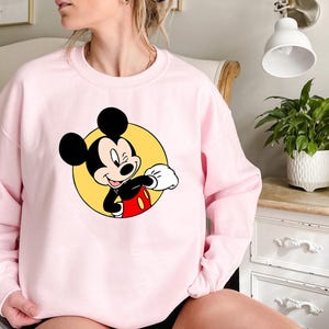 May include: A pink sweatshirt with a graphic of Mickey Mouse in a yellow circle. Mickey Mouse is wearing red shorts and white gloves. He is smiling and looking to the right.