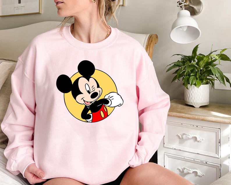 May include: A pink sweatshirt with a graphic of Mickey Mouse in a yellow circle. Mickey Mouse is wearing red shorts and white gloves. He is smiling and looking to the right.