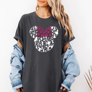 May include: A dark gray t-shirt with a white silhouette of Mickey Mouse's head made up of various Disney characters. The bow on Mickey's head is pink with white polka dots.