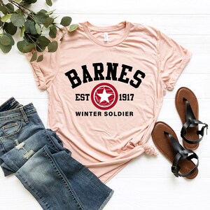 May include: A light pink t-shirt with the word "BARNES" in black block letters above a red and white star emblem. Below the emblem are the words "EST 1917 WINTER SOLDIER". The shirt is paired with blue jeans and brown sandals.