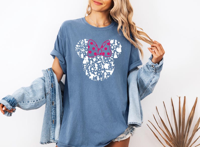 May include: A blue t-shirt with a white silhouette of Mickey Mouse's head made up of various Disney characters. The bow is pink with white polka dots.