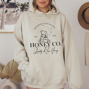 May include: A cream-colored hoodie with a black graphic of Winnie the Pooh holding a jar of honey. The graphic also includes the text "Hundred Acre Woods Est. 1926 Honey Co. Quality Raw Honey".