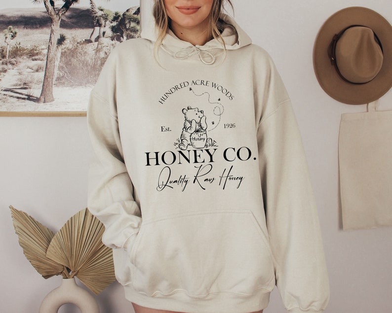 May include: A cream-colored hoodie with a black graphic of Winnie the Pooh holding a jar of honey. The graphic also includes the text "Hundred Acre Woods Est. 1926 Honey Co. Quality Raw Honey".