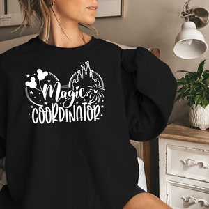 May include: Black sweatshirt with a white graphic of Mickey Mouse ears with the text "Magic Coordinator" inside the ears. The graphic also includes a silhouette of Cinderella's castle and fireworks.