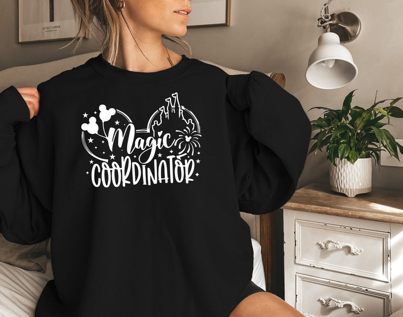 May include: Black sweatshirt with a white graphic of Mickey Mouse ears with the text "Magic Coordinator" inside the ears. The graphic also includes a silhouette of Cinderella's castle and fireworks.